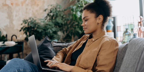 Woman in brown jacket sitting on couch typing on her laptop