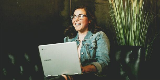 Woman in denim jacket and glasses sitting down holding a laptop